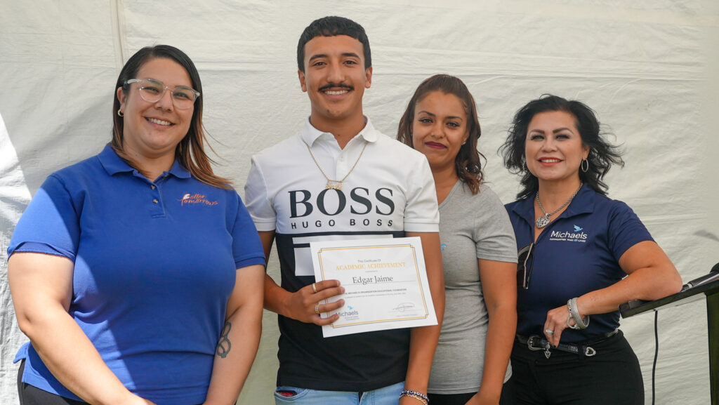 Three people posing with a Michaels Scholar who is holding a certificate of achievement